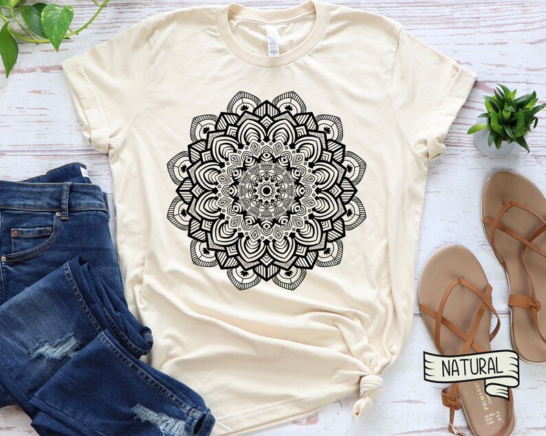 May include: A beige t-shirt with a black mandala design. The shirt is laid flat on a white wooden surface with a pair of brown sandals and a pair of blue jeans.