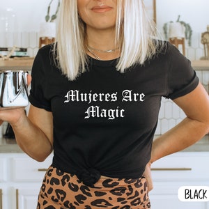 May include: Black t-shirt with the text "Mujeres Are Magic" in white lettering. The shirt is worn by a person with blonde hair and is knotted at the waist.