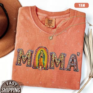 May include: A peach-colored t-shirt with the word "MOMA" printed in colorful letters with floral designs. The letter "O" features an image of the Virgin Mary.
