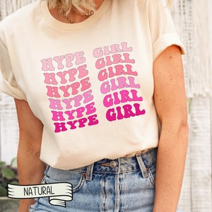 May include: Cream-colored t-shirt with the words "HYPE GIRL" in a repeating pattern of pink and fuchsia bubble letters. The shirt has short sleeves and a relaxed fit. The word "NATURAL" is printed on a banner at the bottom.
