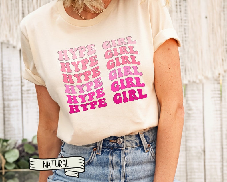 May include: Cream-colored t-shirt with the words "HYPE GIRL" in a repeating pattern of pink and fuchsia bubble letters. The shirt has short sleeves and a relaxed fit. The word "NATURAL" is printed on a banner at the bottom.