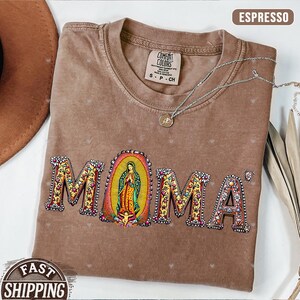 May include: A brown t-shirt with a floral design that spells out "MOMA" with a depiction of the Virgin Mary in the center. The shirt has a distressed look and is made of a soft, comfortable material.
