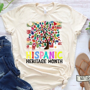 May include: A beige t-shirt with a colorful tree made of handprints. The tree is in front of a distressed American flag with the flags of Latin American countries. The text "HISPANIC HERITAGE MONTH" is below the tree.