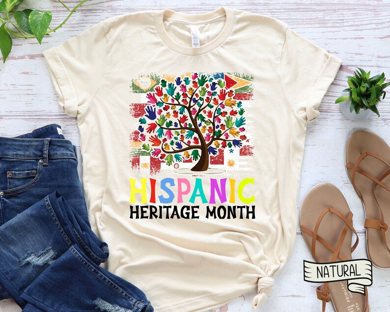 May include: A beige t-shirt with a colorful tree made of handprints. The tree is in front of a distressed American flag with the flags of Latin American countries. The text "HISPANIC HERITAGE MONTH" is below the tree.