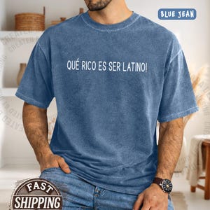 May include: Blue jean-colored t-shirt with the text "QUÉ RICO ES SER LATINO!" in white. The shirt has a crew neck and short sleeves. The words "BLUE JEAN" are printed in a blue rectangle in the upper right corner.