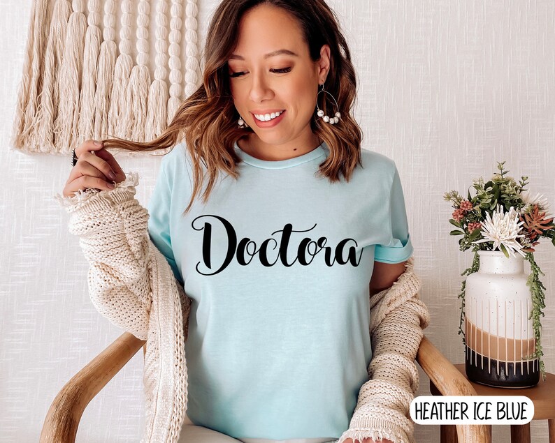 K&ouml;nnte beinhalten: Ein hellblaues T-Shirt mit dem Wort "Doctora" in schwarzer, kursiv geschriebener Schrift. Das Shirt wird von einer Person mit langen braunen Haaren getragen und wird mit einem wei&szlig;en Cardigan kombiniert.