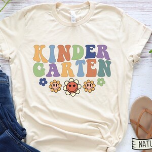 Puede incluir: Una camiseta beige con el texto "KINDERGARTEN" en colores arco&iacute;ris con una fuente retro. Tres flores amarillas con caras sonrientes y centros rojos est&aacute;n debajo del texto.