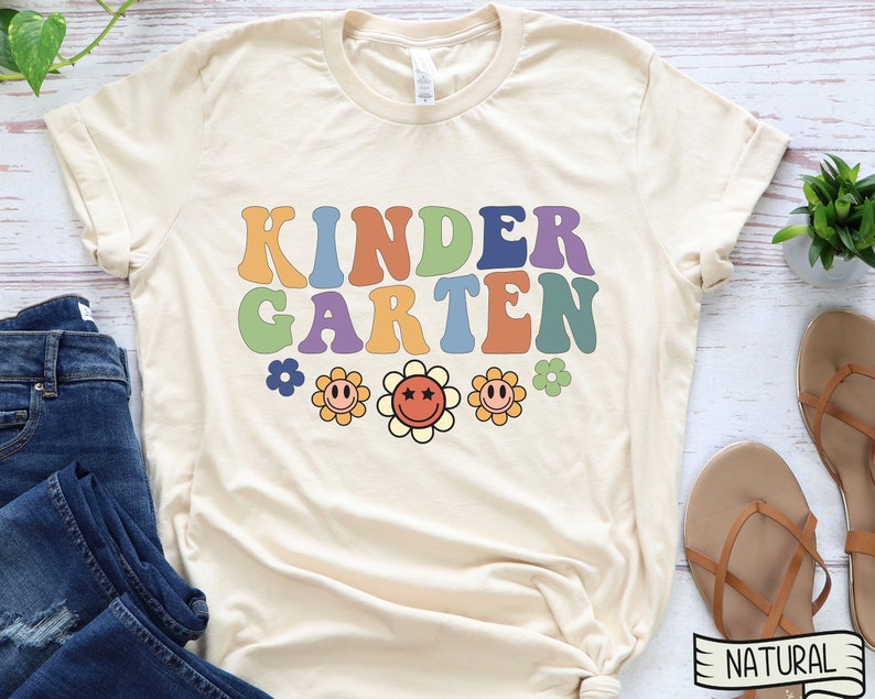 Puede incluir: Una camiseta beige con el texto "KINDERGARTEN" en colores arco&iacute;ris con una fuente retro. Tres flores amarillas con caras sonrientes y centros rojos est&aacute;n debajo del texto.