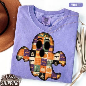May include: A violet Comfort Colors t-shirt featuring a patchwork ghost design with pumpkins, bats, and haunted houses. The ghost has black eyes and mouth. A gold necklace is draped across the shirt. The shirt has a small Comfort Colors tag.