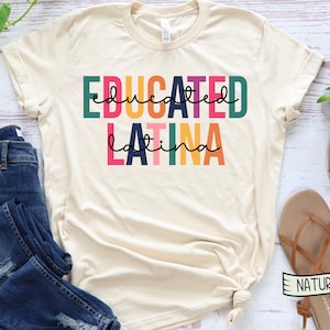 May include: A beige t-shirt with the text "educated latina" in colorful block letters. The shirt is laid flat on a white wooden surface with a pair of brown sandals and blue jeans.