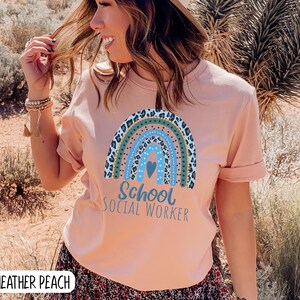 Puede incluir: Una camiseta rosa con un dise&ntilde;o de arco&iacute;ris con estampado de leopardo, lunares y un coraz&oacute;n. El texto "School Social Worker" est&aacute; impreso debajo del arco&iacute;ris.