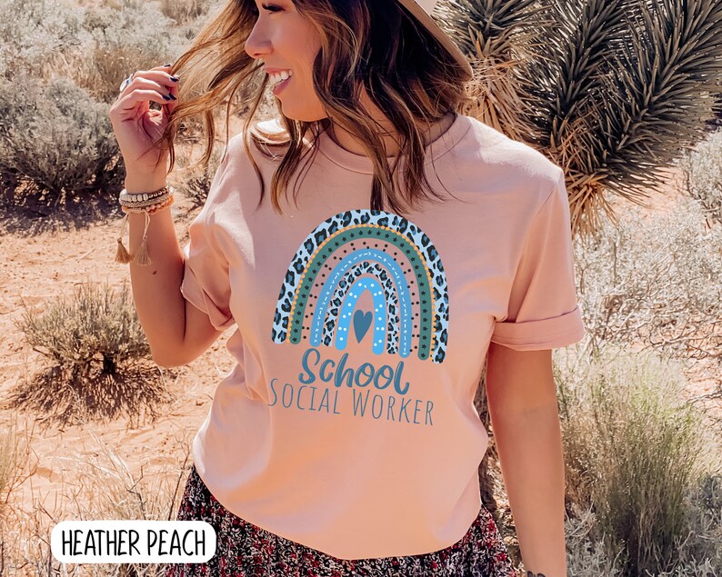 Puede incluir: Una camiseta rosa con un dise&ntilde;o de arco&iacute;ris con estampado de leopardo, lunares y un coraz&oacute;n. El texto "School Social Worker" est&aacute; impreso debajo del arco&iacute;ris.