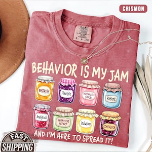 May include: Crimson Comfort Colors t-shirt with the phrase "BEHAVIOR IS MY JAM" and illustrated jars labeled with positive attributes. The shirt also includes the text "AND I'M HERE TO SPREAD IT!" A silver necklace is draped across the shirt.