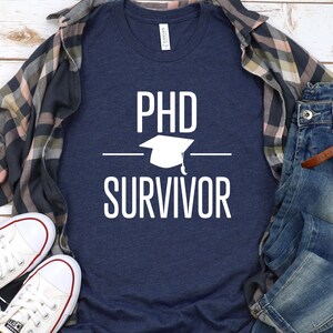 May include: A navy blue t-shirt with white text that reads "PhD Survivor" and a graduation cap graphic.