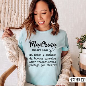 May include: A light blue t-shirt with the word "Madrina" in black text, defined as "[madre-nah]" and the text "da besos y abrazos, da buenos consejos, amor incondicional, protege por siempre". The shirt is on a person wearing it. The text "HEATHER ICE BLUE" is in the bottom right corner of the image.