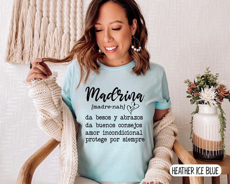 May include: A light blue t-shirt with the word "Madrina" in black text, defined as "[madre-nah]" and the text "da besos y abrazos, da buenos consejos, amor incondicional, protege por siempre". The shirt is on a person wearing it. The text "HEATHER ICE BLUE" is in the bottom right corner of the image.