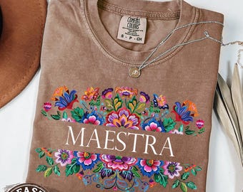 May include: Espresso-colored t-shirt with the word "MAESTRA" in white, framed by a colorful floral design. The shirt has a crew neck and short sleeves. A silver necklace with a pendant is draped across the shirt.