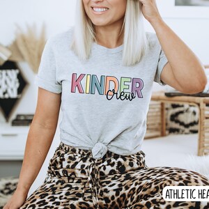 May include: A gray t-shirt with the text "Kinder Crew" in rainbow colors. The shirt is tied at the waist and the model is wearing leopard print pants. The text "Athletic Heather" is visible in the bottom right corner.