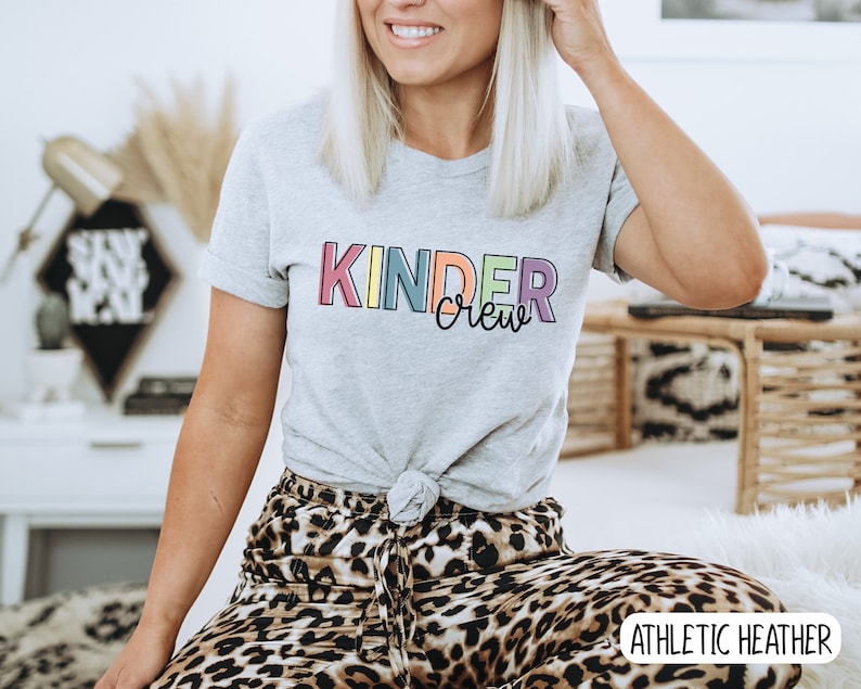 May include: A gray t-shirt with the text "Kinder Crew" in rainbow colors. The shirt is tied at the waist and the model is wearing leopard print pants. The text "Athletic Heather" is visible in the bottom right corner.