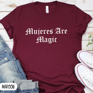May include: A maroon t-shirt with the text "Mujeres Are Magic" in white lettering. The shirt is laid flat on a wooden surface with a pair of white sneakers and blue jeans.