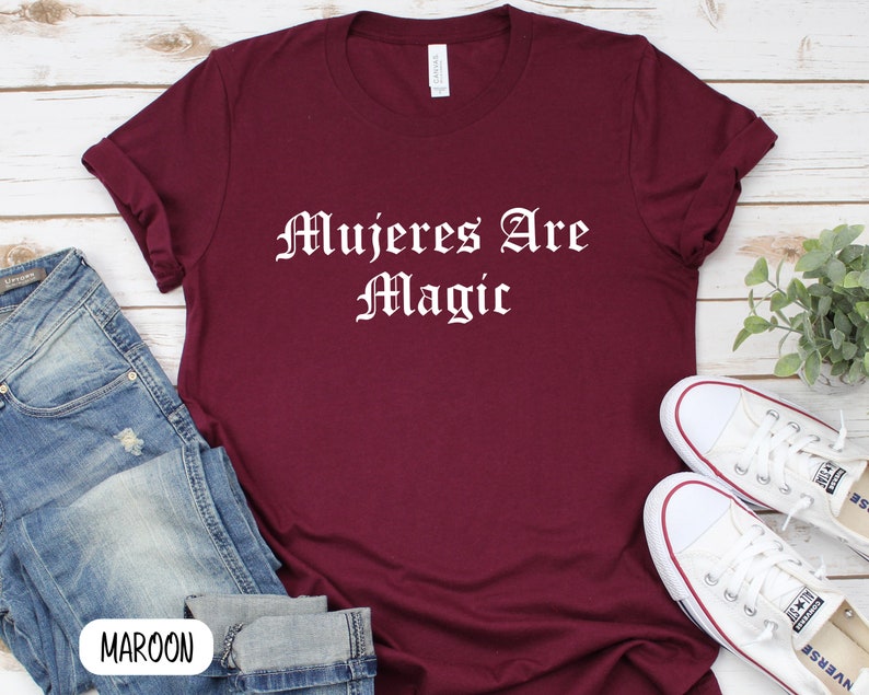 May include: A maroon t-shirt with the text "Mujeres Are Magic" in white lettering. The shirt is laid flat on a wooden surface with a pair of white sneakers and blue jeans.