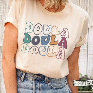 May include: A cream colored t-shirt with the word "Doula" repeated three times in a retro font with different colors. The shirt is being worn by a person with blue jeans. The text "Soft Cream" is visible in the lower right corner of the image.