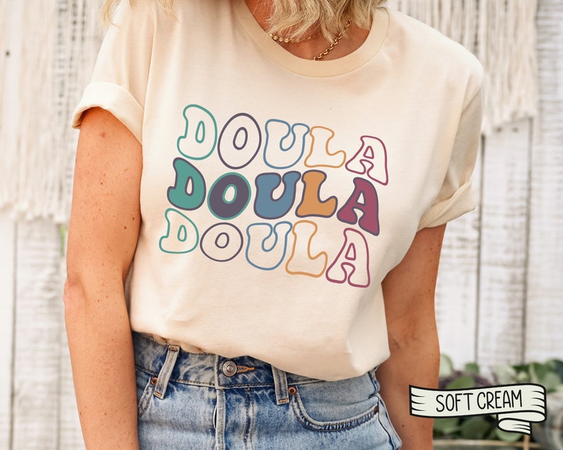 May include: A cream colored t-shirt with the word "Doula" repeated three times in a retro font with different colors. The shirt is being worn by a person with blue jeans. The text "Soft Cream" is visible in the lower right corner of the image.