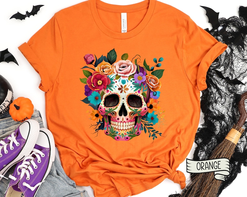 May include: Orange t-shirt with a colorful sugar skull design surrounded by flowers. The skull has intricate details and is surrounded by a variety of flowers in shades of pink, purple, blue, and yellow. The t-shirt has the word 'ORANGE' written on a tag in the bottom right corner.