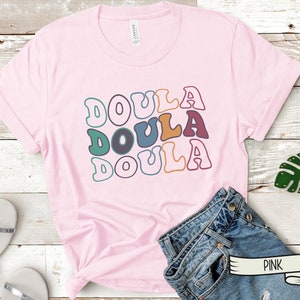 May include: Pink t-shirt with the word "Doula" repeated three times in a retro font with different colors. The shirt is laying on a wooden surface with a pair of jeans and a green leaf.