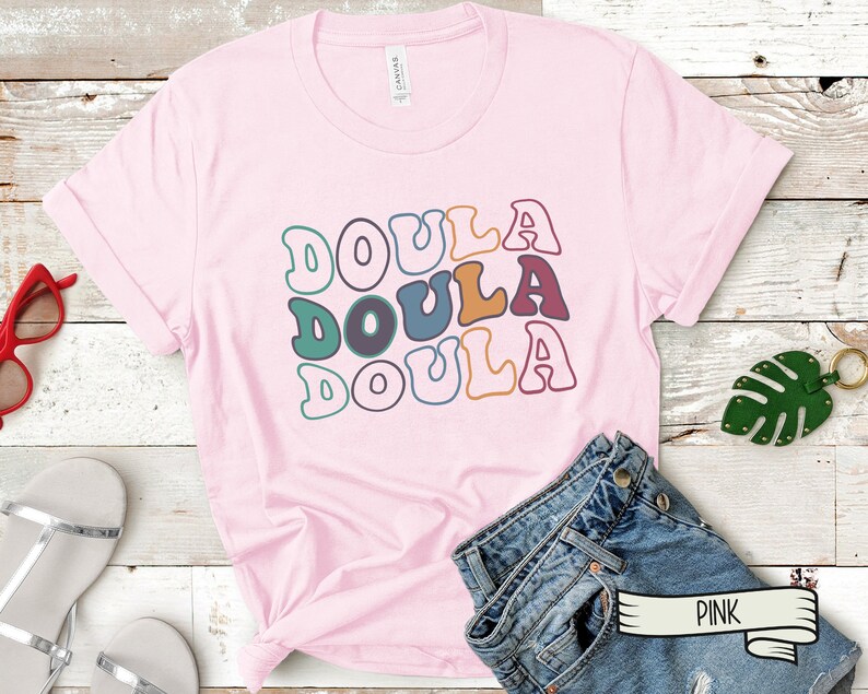 May include: Pink t-shirt with the word "Doula" repeated three times in a retro font with different colors. The shirt is laying on a wooden surface with a pair of jeans and a green leaf.