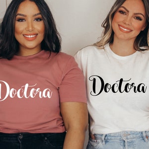 K&ouml;nnte beinhalten: Zwei Personen tragen T-Shirts mit dem Wort "Doctora" in kursivschrift. Ein Shirt ist rosa und das andere wei&szlig;.