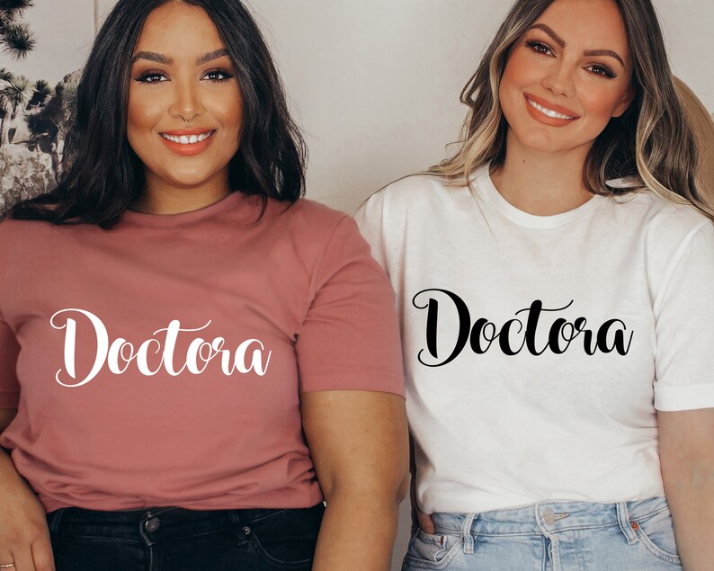 K&ouml;nnte beinhalten: Zwei Personen tragen T-Shirts mit dem Wort "Doctora" in kursivschrift. Ein Shirt ist rosa und das andere wei&szlig;.