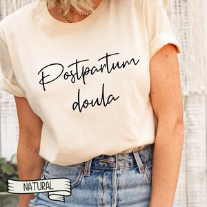 May include: A white t-shirt with the text "Postpartum Doula" in black script lettering. The shirt is being worn by a person with blue jeans and a gold necklace. The word "NATURAL" is printed on a white ribbon.