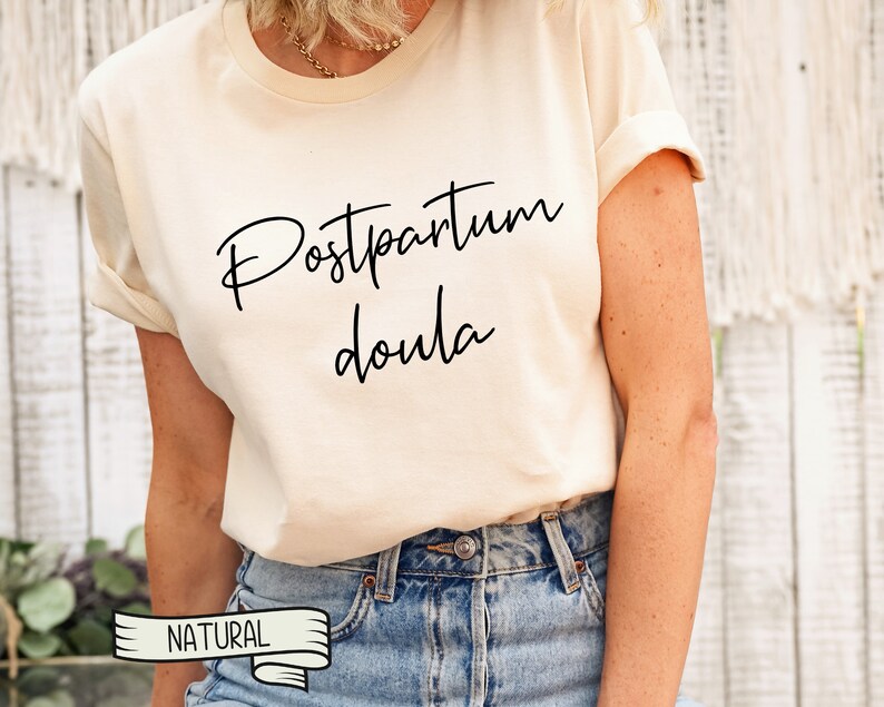 May include: A white t-shirt with the text "Postpartum Doula" in black script lettering. The shirt is being worn by a person with blue jeans and a gold necklace. The word "NATURAL" is printed on a white ribbon.