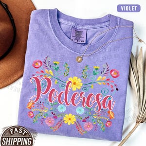 May include: A violet Comfort Colors t-shirt with the word "Poderosa" in pink script, surrounded by a floral design with yellow, pink, and blue flowers. A gold necklace rests on the shirt.