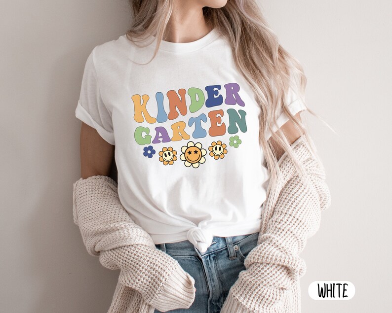 Puede incluir: Camiseta blanca con el texto "KINDERGARTEN" en letras retro de colores y una fila de caras de flores sonrientes.