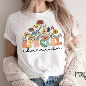 May include: White t-shirt featuring a floral design with the words "SPECIAL education" in colorful, retro-style lettering. The flowers include yellow, orange, and red blooms. The shirt is a classic crew neck.