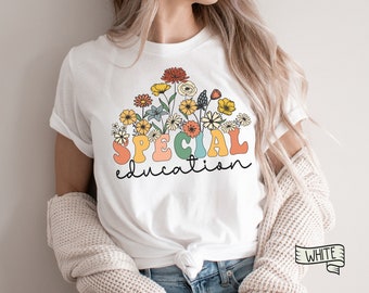 May include: White t-shirt featuring a floral design with the words "SPECIAL education" in colorful, retro-style lettering. The flowers include yellow, orange, and red blooms. The shirt is a classic crew neck.