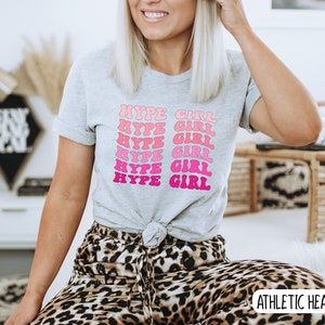 May include: A light gray athletic heather t-shirt with the text "HYPE GIRL" in a repeating, wavy design of pink and fuchsia. The model is wearing leopard print pants.