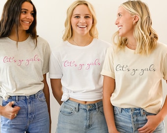 May include: Three women wearing t-shirts with the text "Let's go girls" in different colors. The shirts are white, grey, and beige.