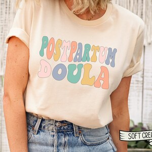 May include: A cream colored t-shirt with the text "POSTPARTUM DOULA" in rainbow colored letters. The text is in a retro font style. The t-shirt is being worn by a person. The text "SOFT CREAM" is visible in the lower right corner of the image.