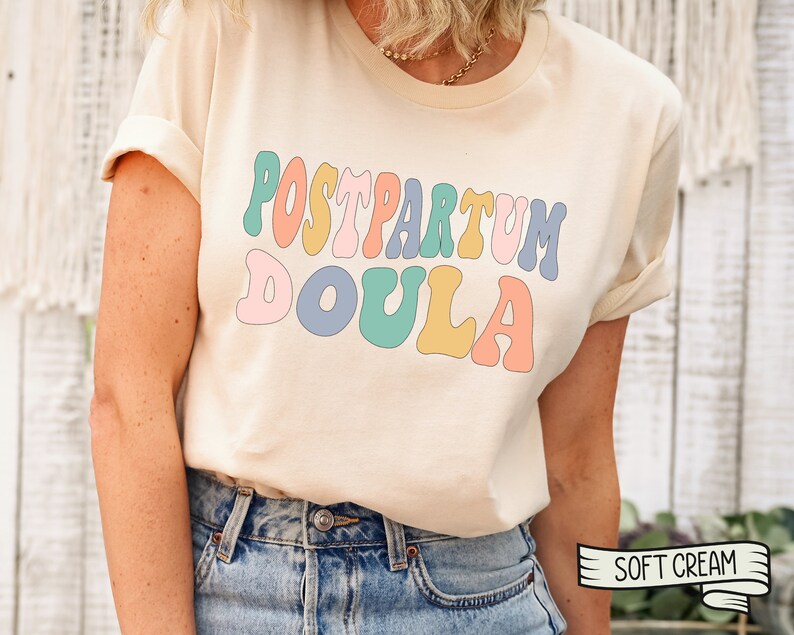 May include: A cream colored t-shirt with the text "POSTPARTUM DOULA" in rainbow colored letters. The text is in a retro font style. The t-shirt is being worn by a person. The text "SOFT CREAM" is visible in the lower right corner of the image.