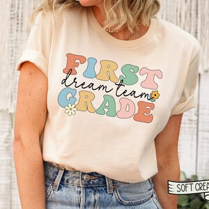 Op de afbeelding: Een licht beige T-shirt met de tekst "FIRST dream team GRADE" in kleurrijke, retro-stijl letters. De letters zijn zwart omrand en hebben een licht reli&euml;feffect. Er zijn kleine bloemenillustraties naast de tekst.