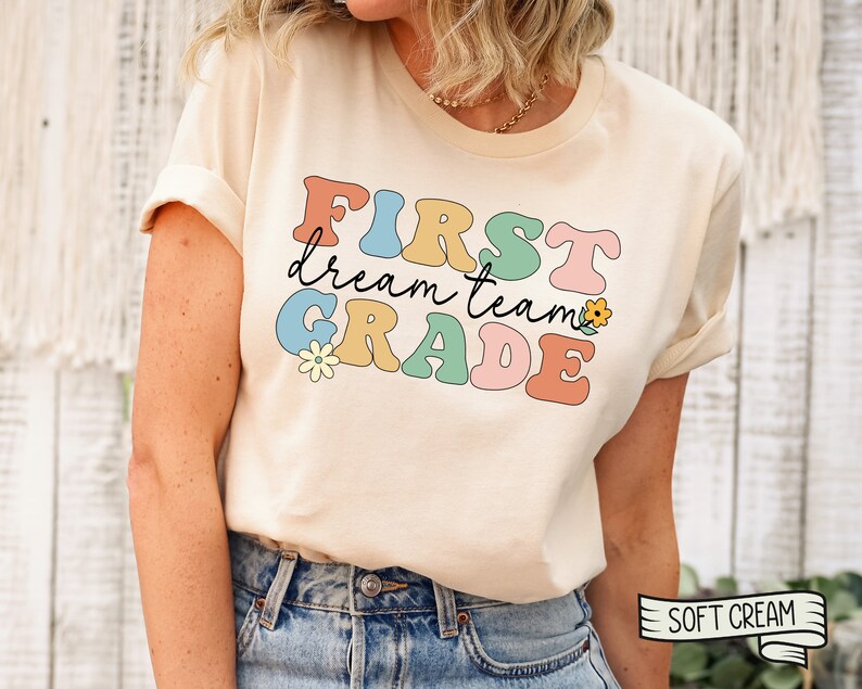 Op de afbeelding: Een licht beige T-shirt met de tekst "FIRST dream team GRADE" in kleurrijke, retro-stijl letters. De letters zijn zwart omrand en hebben een licht reli&euml;feffect. Er zijn kleine bloemenillustraties naast de tekst.