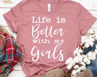 以下が含まれることがあります： 白地に「Life is Better with my Girls」と書いてある、くすんだピンクのTシャツです。
