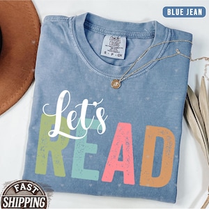 May include: Blue jean-colored t-shirt with the words "Let's READ" in colorful letters. The shirt has a crew neck and short sleeves. A gold necklace with a pendant is draped across the shirt. The shirt is folded on a white surface.
