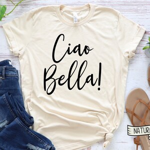 K&ouml;nnte beinhalten: Cremefarbenes T-Shirt mit dem Schriftzug "Ciao Bella!" in schwarzer Schrift. Das Shirt wird mit blauen Jeans und braunen Sandalen kombiniert. Das Wort "NATURAL" ist auf einem kleinen Etikett aufgedruckt.