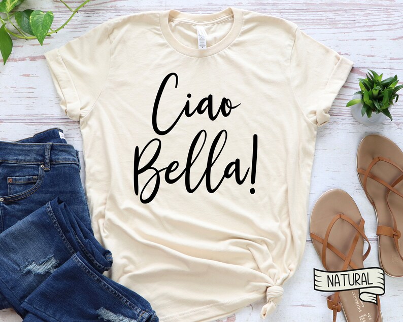 K&ouml;nnte beinhalten: Cremefarbenes T-Shirt mit dem Schriftzug "Ciao Bella!" in schwarzer Schrift. Das Shirt wird mit blauen Jeans und braunen Sandalen kombiniert. Das Wort "NATURAL" ist auf einem kleinen Etikett aufgedruckt.