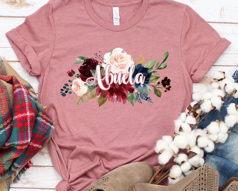 Pu&ograve; includere: Una maglietta color malva con un disegno floreale e la parola "Abuela" in corsivo rosa.