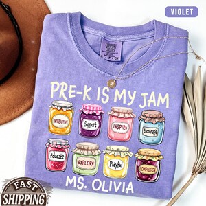 May include: Purple t-shirt with the text "PRE-K IS MY JAM" and a row of jam jars with words like "Interactive", "Support", "Inspire", "Encourage", "Educate", "Explore", "Playful", and "Compassion". The text "MS. OLIVIA" is below the jars.